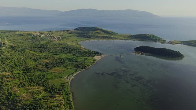 Aerial view of the Vjose-Narte Delta Protected Area featuring Zvernec Island and its wooden bridge surrounded by lush hills and calm lagoon waters in Zvernec, Vlore County, Albania.
