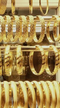 Rows of shiny gold bangles and bracelets hang in a jewelry shop display case as the camera holds on the ornate metal designs and reflections