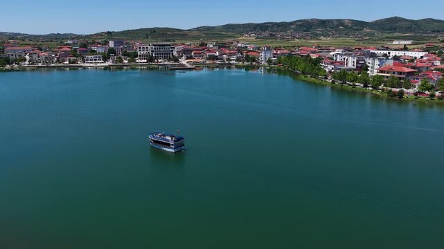 Aerial view of a passenger boat cruising on the calm waters of Lake Seferan with the town of Belsh and rolling green hills in the background, Belsh, Elbasan County, Albania.