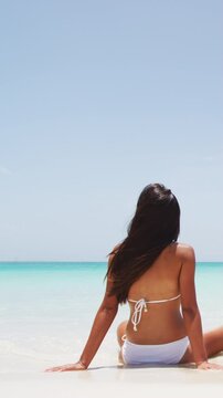 Vertical video. Beach vacation dream woman enjoying summer holiday on dreamy perfect ocean tropical destination. Person sitting from the back alone on deserted white sand beach getaway. Copy space.