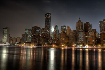 Fototapeta premium New York City skyline at night with Midtown Manhattan skyscrapers and the illuminated Chrysler Building reflecting across the East River seen from Roosevelt Island waterfront overlook