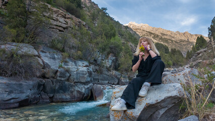 Naklejka premium Sitting peacefully by a shimmering river, a young woman enjoys the tranquility of the surrounding mountains. She holds a bouquet of bright flowers, embracing the calm afternoon light.