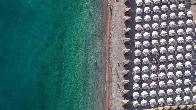 Aerial view of rows of white beach umbrellas on the sandy shore of Llamani Beach with turquoise water and people swimming, Himare, Vlore County, Albania.