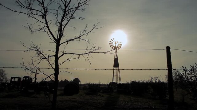 Windmill Silhouette Spinning Against Sun With Tractor Moving In Farm - 4K