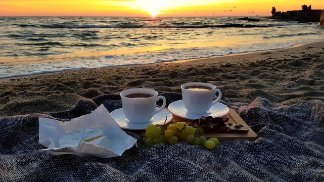 Luxury romantic beach picnic at sunset. The frame features two white cups, fresh green grapes, chocolate, and an open wheel of gourmet brie cheese on blanket against the background of calm sea waves.