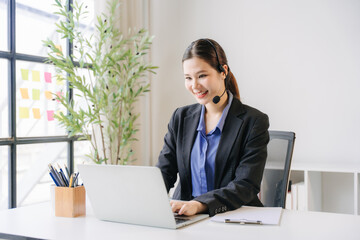 Fototapeta premium A Cheerful Call Center Agent at Work: A smiling young woman wearing a headset, dressed in professional attire, is actively engaged on her laptop in a bright office setting.
