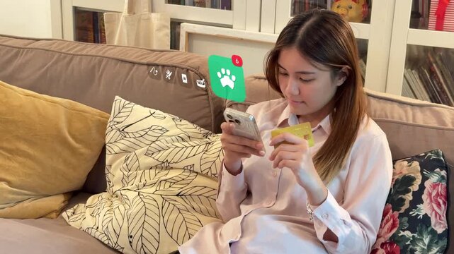 Living room shot of Southeast Asian woman shopping for pets on mobile with credit card, featuring a 3D tracked animal paw badge and cat litter box icons.
