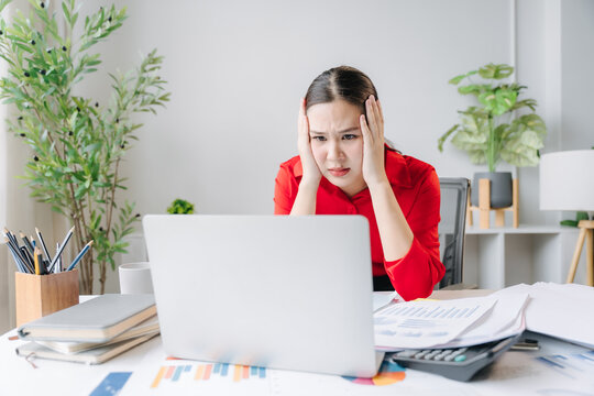 Stressful Office Moment: A young woman in a red blouse clutches her head in frustration as she stares at a laptop screen, overwhelmed by tasks and deadlines in a cluttered workspace.