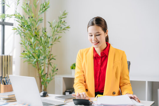 Smiling Businesswoman Calculating Finances: A young, cheerful businesswoman with an Asian ethnicity, wearing a bright yellow blazer over a red shirt, sits at her desk using a calculator and laptop.