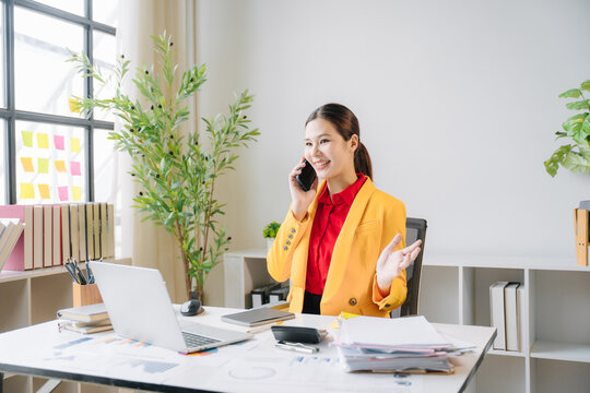 Focused Professional at Work: A vibrant businesswoman in a yellow blazer smiles as she engages in a phone conversation, her workspace filled with organized documents, a laptop, and lush green plants.