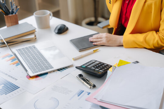 Deskwork Elegance: A woman's hands busy at a desk, surrounded by charts, a laptop, and a calculator, epitomizing focused productivity and professional diligence.