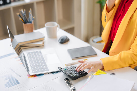 Financial Focus: A woman in a vibrant yellow blazer intently uses a calculator at her desk, surrounded by financial documents and a laptop, signifying meticulous financial planning and analysis.