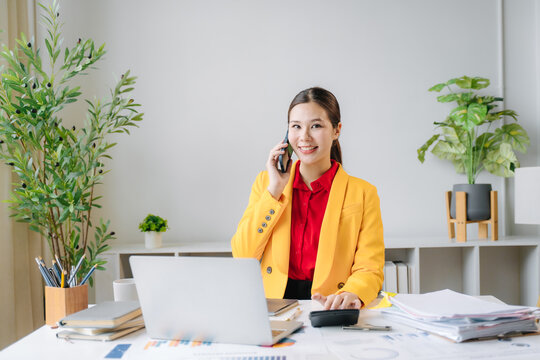 Professional Woman in Yellow Jacket on Phone: A young businesswoman in a vibrant yellow jacket and red shirt smiles while talking on the phone at her desk, surrounded by plants, a laptop.