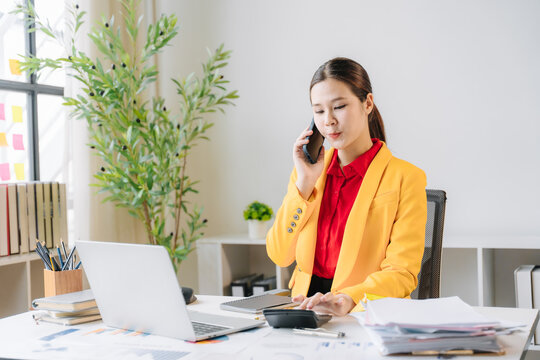 Businesswoman on the Phone: A focused businesswoman in a yellow blazer is working at her desk, using a calculator and speaking on her mobile phone, surrounded by documents and office supplies.