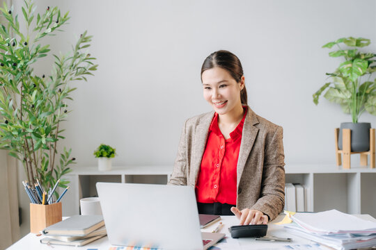Focused Professional at Work: A smiling businesswoman in a smart blazer and red shirt works diligently at her desk, illuminated by the natural light of an office.