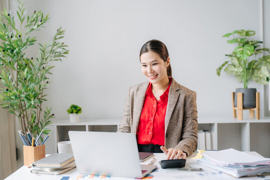Focused Professional at Work: A young Asian businesswoman smiles while working on her laptop and calculator at her desk, surrounded by plants and documents, signifying a productive office environment.