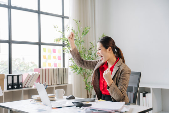 Celebration of Success: A jubilant businesswoman exclaims with joy, fists clenched in a triumphant gesture, as she looks at her computer screen in a bright, modern office setting.