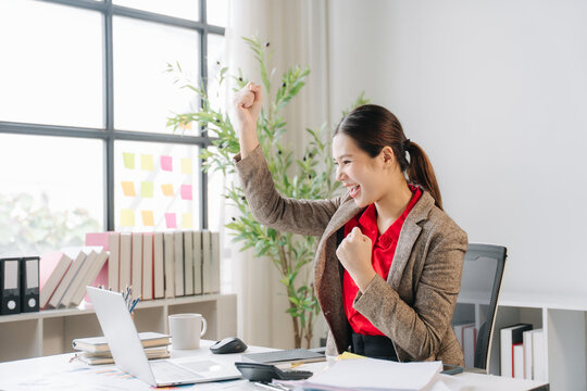 Excited businesswoman celebrating success in office: A young businesswoman smiles with joy and triumph as she raises her fist in a celebration of achievement at her modern office desk.