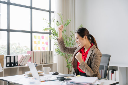 Triumph at Desk: A woman in a business suit celebrates a successful outcome at her office desk, raising her fist in a gesture of accomplishment. Surrounded by office supplies and natural light.