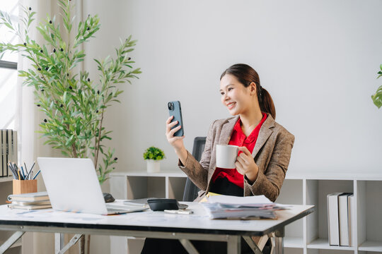 Video Call Comfort: A woman in business attire smiles while holding a mug and looking at her phone, perhaps on a video call, in a bright office setting with a laptop and plant.