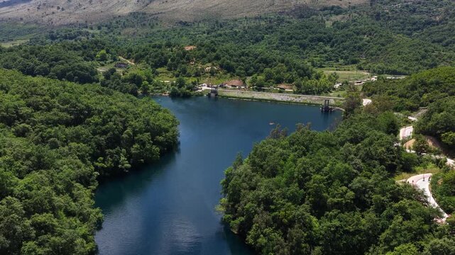Aerial view of the Blue Eye natural spring surrounded by dense green forest and rolling hills under a clear sky in Finiq, Vlore County, Albania.