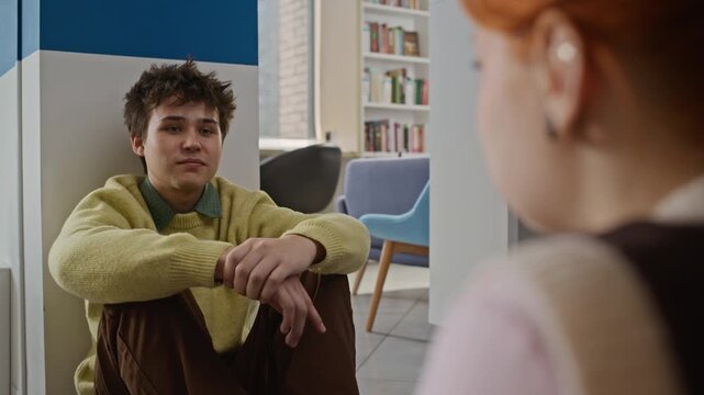 Over shoulder shot of Caucasian teenage boy sitting on floor and talking to female classmate in library. Video for educational marketing, student life promotions, or peer-to-peer learning campaigns