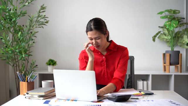 Stressful Office Work: A woman in a red shirt, overwhelmed by work, sits at her desk with a laptop, calculator, and papers, looking stressed and wiping tears from her eyes.