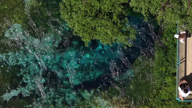 Aerial view of the Blue Eye natural spring with crystal clear turquoise water and people on a wooden platform surrounded by lush green trees in Finiq, Vlore County, Albania.