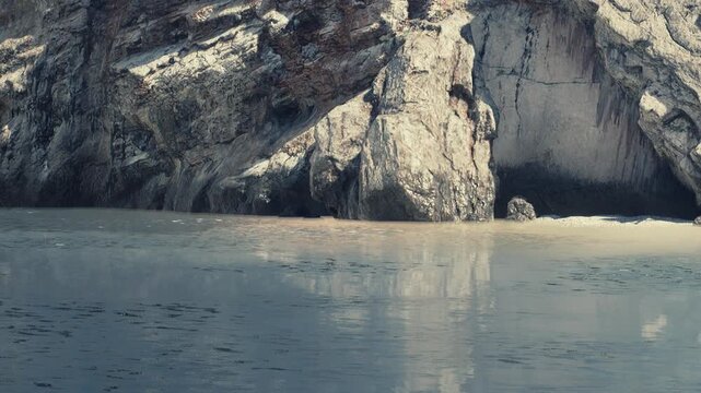 Rocks stand tall beside clear water with sand visible at the shore. The setting shows a coastal area where land meets sea. Light plays on the surface of the water.