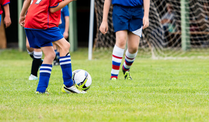 Fototapeta premium Young players in red and blue converge near a goal net, driving the ball into the attacking third where a youth match is most likely to be decided. The netting behind them frames the action