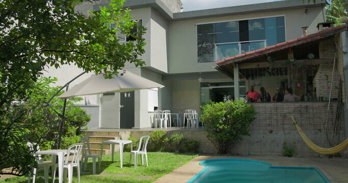 Sunny backyard scene with umbrella-shaded tables, hammock, swimming pool, and group socializing at covered grill area of modern suburban home