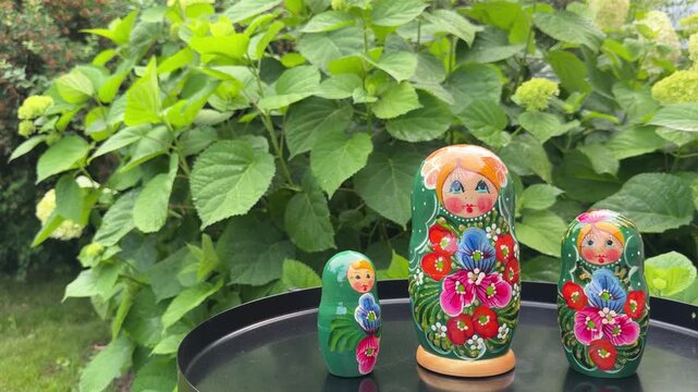 Russian Nesting Dolls on a Table: Three vibrant Russian nesting dolls, adorned with floral patterns and cheerful faces, stand on a dark round table against a backdrop of lush green foliage.