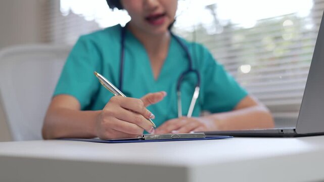 Young Asian female doctor in green scrubs with a stethoscope providing medical advice to a patient through a video call, carefully taking notes on a clipboard in her modern clinic office
