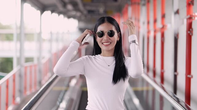 Over-the-shoulder shot of a cheerful young Thai woman wearing trendy sunglasses. She turns around, smiles brightly at the camera, and walks confidently on an urban skywalk at a public transit station.
