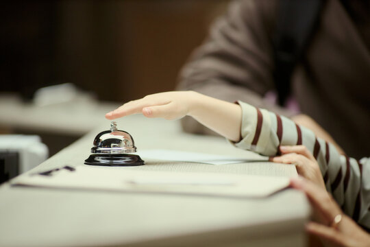 Close up of child hand reaching and pressing hotel reception bell while family checking in at front desk. Ideal for travel booking, hospitality service, family vacation marketing