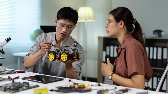 Asian male engineer assembling a wheeled robot and explaining its mechanics to a female colleague in a modern technology lab, collaborating on a STEM electronics project together