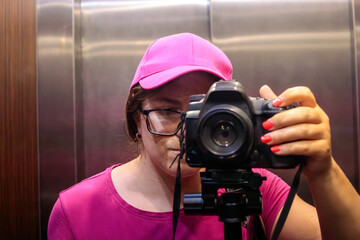Woman photographer in pink cap and glasses taking mirror selfie with DSLR camera on tripod inside elevator © Татьяна Швец
