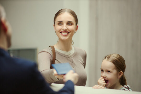 Caucasian mother checking in at hotel reception with child, handing over passport while speaking with staff. Travel accommodation, family tourism, hospitality marketing concept, close up