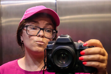 Woman photographer in pink cap and glasses making pouty face taking mirror selfie with DSLR camera in elevator © Татьяна Швец