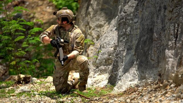 Camouflaged soldier kneels beside a rocky cliff, aiming a rifle during a tactical operation in rugged outdoor terrain.