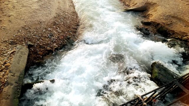 Fast-moving water churns through a narrow rocky channel, foaming between sandy banks and broken concrete, resembling a drainage spillway or stream