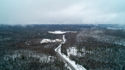 Fototapeta premium A sweeping high-angle drone photograph over a winding two-lane highway cutting through a dense winter forest, with low hanging fog and mist drifting below a moody, overcast sky.