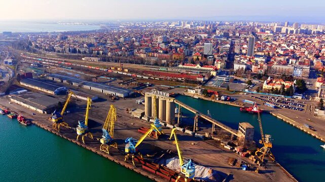 Elevated view of a commercial seaport and city. Drone panorama showing yellow dock crane, railway track, industrial warehouse and dense urban building under clear sky.