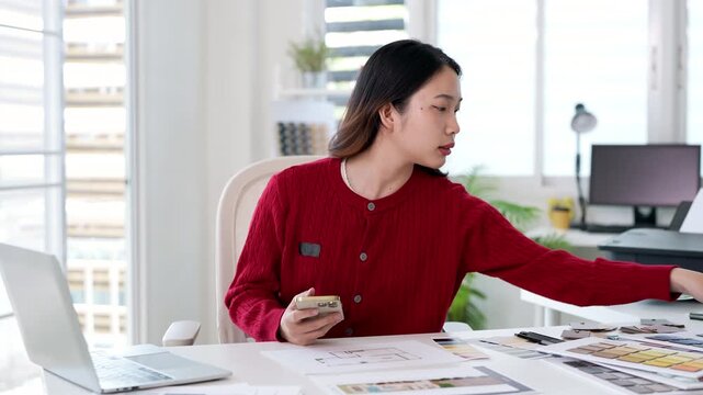 Concentrated young interior designer woman sitting at table with blueprint drawing ,swatches and material , typing on phones researching information about materials for decorations.
