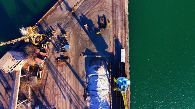 Harbor crane and covered cargo storage area, Burgas. Drone view of a yellow industrial lifter casting a long shadow next to a mound and train track.
