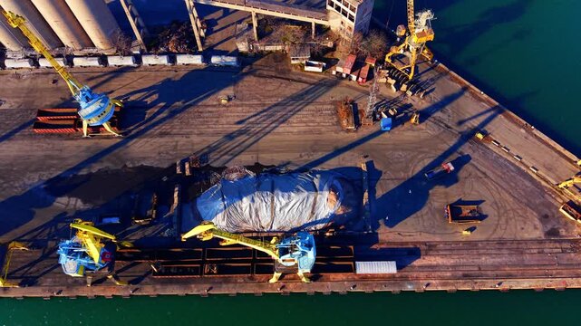 Overhead view of a dock crane and cargo pile. Yellow machine casting a long shadow on concrete ground next to a covered material heap.