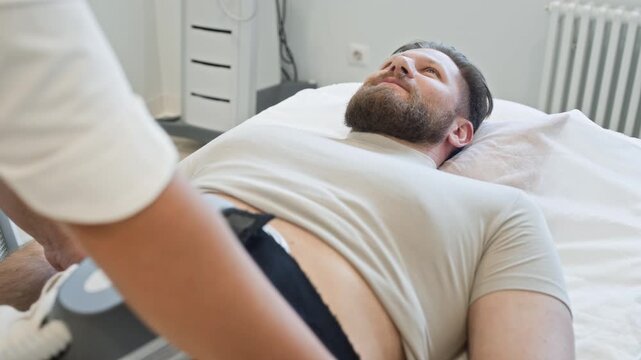 Caucasian man lying on clinic bed while practitioner adjusting muscle stimulation device on abdomen. Ideal for promoting non-invasive body sculpting treatments