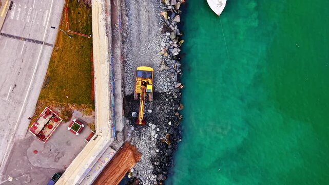 Overhead view of an excavator on a breakwater. Yellow construction machine placing rock and stone along turquoise sea water.