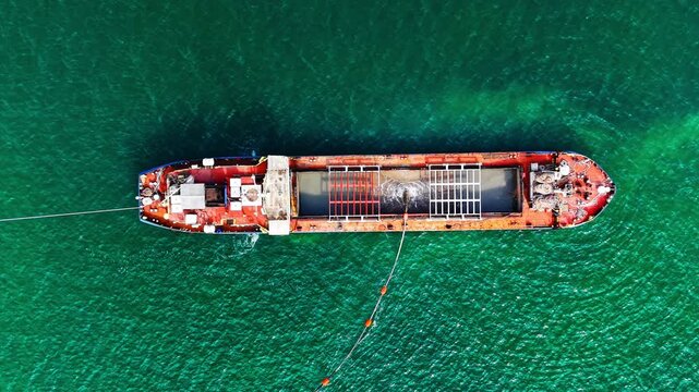 Red dredging barge anchored in turquoise sea. Aerial view of rust red ship hull with open hold and grid framework, mooring lines extending into calm green water.