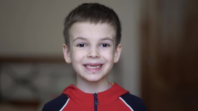 Close-up video of cute young boy showing teeth at camera in red hoodie. Child with short brown hair and open mouth expression in indoor portrait.